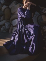 Model wearing a tiered purple dress seated outdoors in front of a stone wall for the Merlette Pre Fall 2025 campaign.