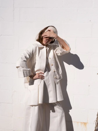 Model Atherton Grace Johnson wearing an off-white buttoned jacket and matching trousers posing against a painted cinder block wall.