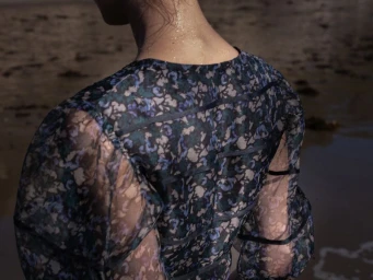 Rear view of a model wearing a sheer blue floral print top outdoors near water for Merlette Pre Fall 2025 Campaign