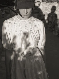 Atherton Grace Johnson in a white lace-trimmed tiered dress and dark brimmed cap under dappled natural sunlight and shadows.