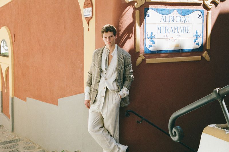 Caesar van den Idsert leans against a terracotta wall wearing a plaid blazer and relaxed linen suit outside Albergo Miramare.