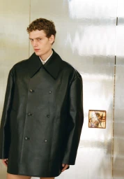 Model Caesar van den Idsert wearing an oversized black leather double-breasted coat against a reflective metallic wall.