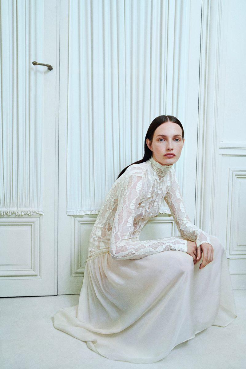 A woman in a lace top and long skirt sits in a white room, with a white door and white curtain visible in the background.