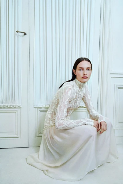 A woman in a lace top and long skirt sits in a white room, with a white door and white curtain visible in the background.