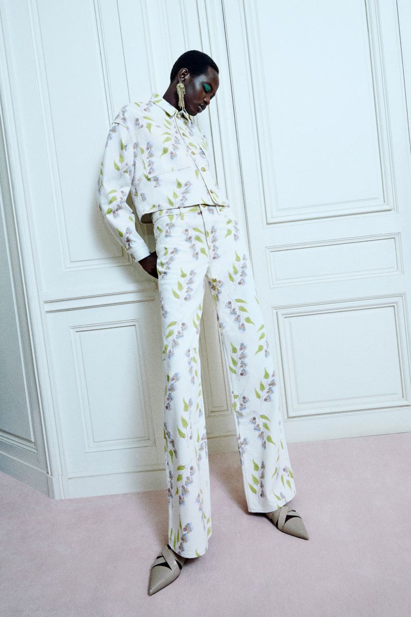 Woman in floral jacket and pantsuit poses against a white paneled wall and pink floor. She wears large gold earrings and taupe heels.