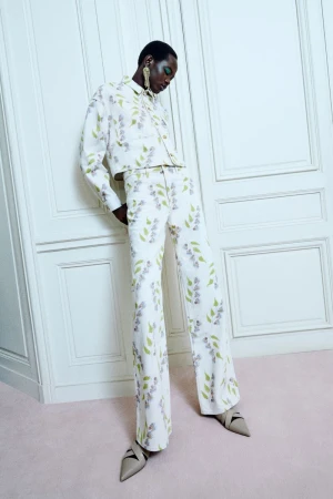 Woman in floral jacket and pantsuit poses against a white paneled wall and pink floor. She wears large gold earrings and taupe heels.