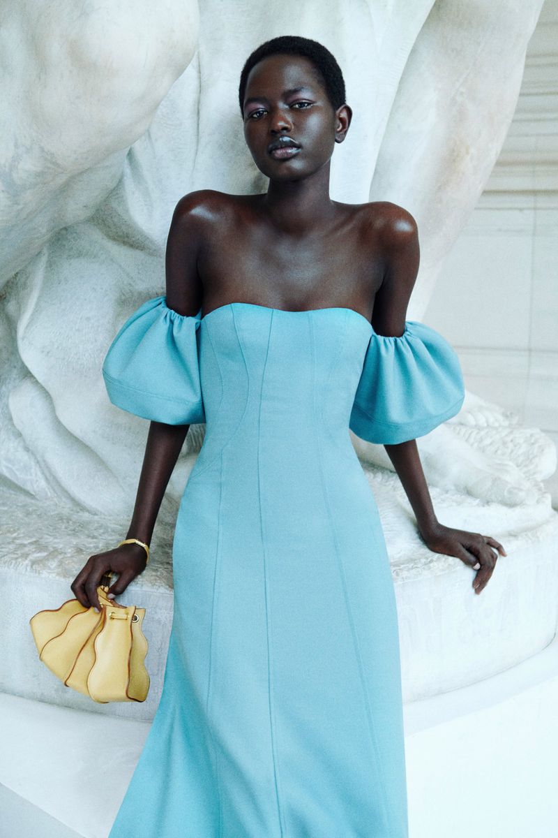 Fashion shoot of a Black woman wearing a light blue gown with puffed sleeves, holding a yellow purse, posing against a white statue.
