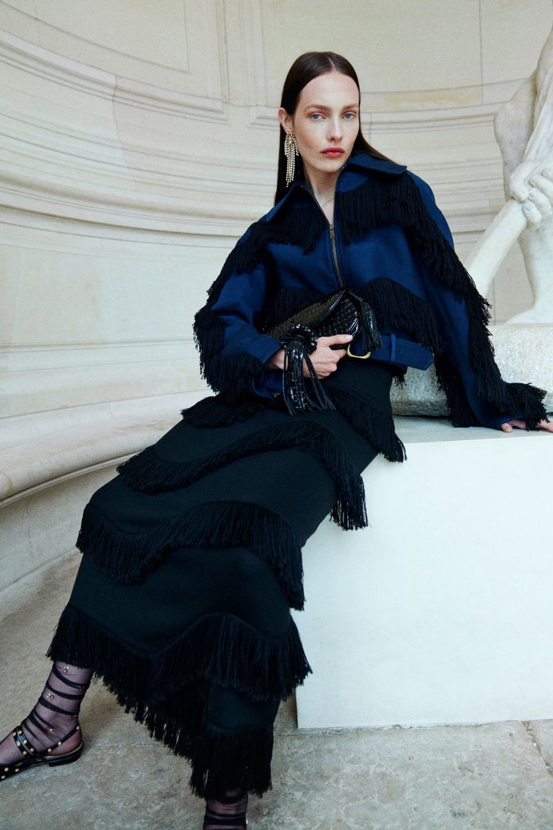 Fashion editorial shot of an adult woman posing in a blue fringe jacket and black fringe skirt, sitting in a stone architectural space.