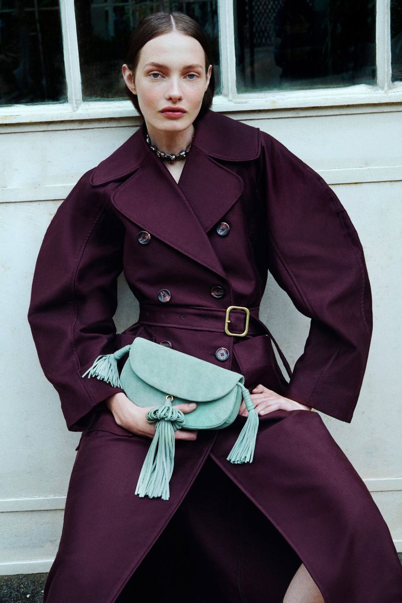 Model Clara Denison in a belted burgundy trench coat holding a mint green tassel clutch against a pale paneled wall.