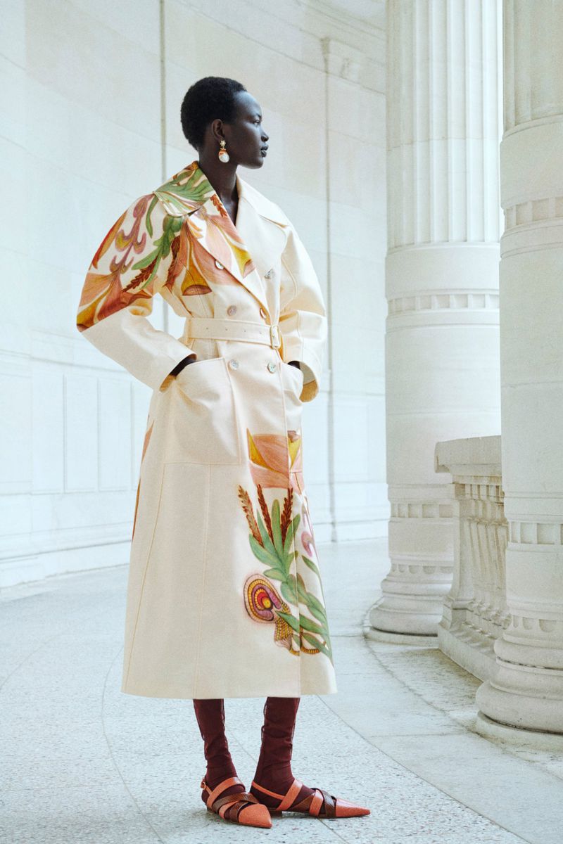 Elegant African-American woman in a floral trench coat and open-toe heels stands amid classical white columns, side profile. Fashion editorial.