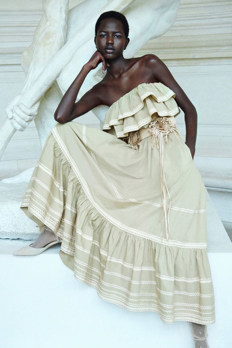 A dark-skinned woman sits by a white marble statue wearing a beige off-the-shoulder dress with a corset and matching pointed-toe pumps.