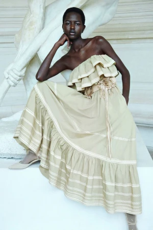 A dark-skinned woman sits by a white marble statue wearing a beige off-the-shoulder dress with a corset and matching pointed-toe pumps.