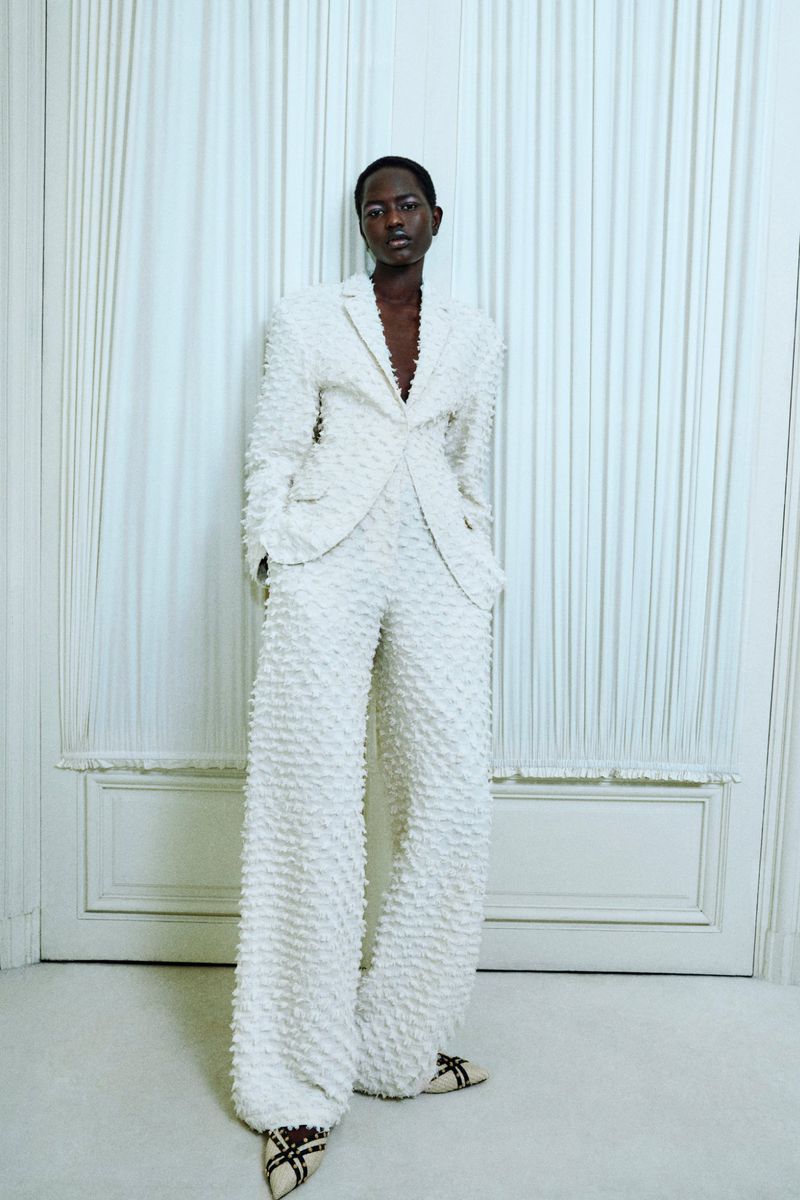 An adult woman in a textured white suit stands indoors in front of a white wall in a fashion shot with studio lighting.