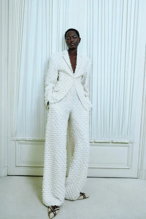 An adult woman in a textured white suit stands indoors in front of a white wall in a fashion shot with studio lighting.