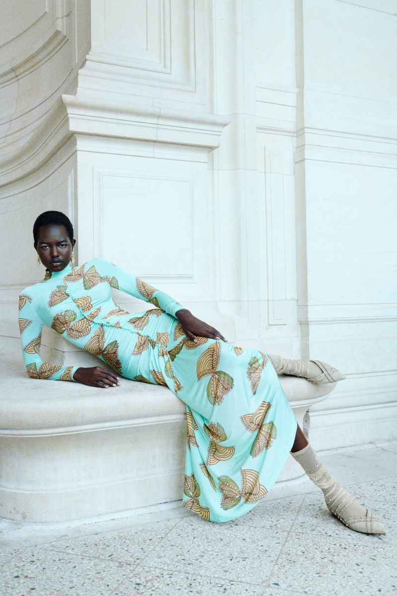 A dark-skinned woman poses while reclining against a white marble wall and base, wearing a patterned, light-blue dress and tall beige sock-boots.