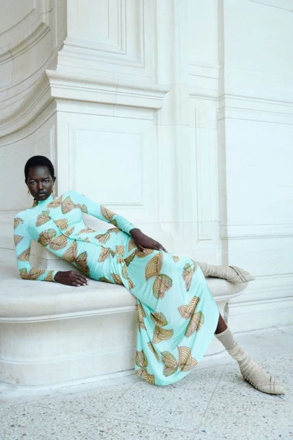 A dark-skinned woman poses while reclining against a white marble wall and base, wearing a patterned, light-blue dress and tall beige sock-boots.