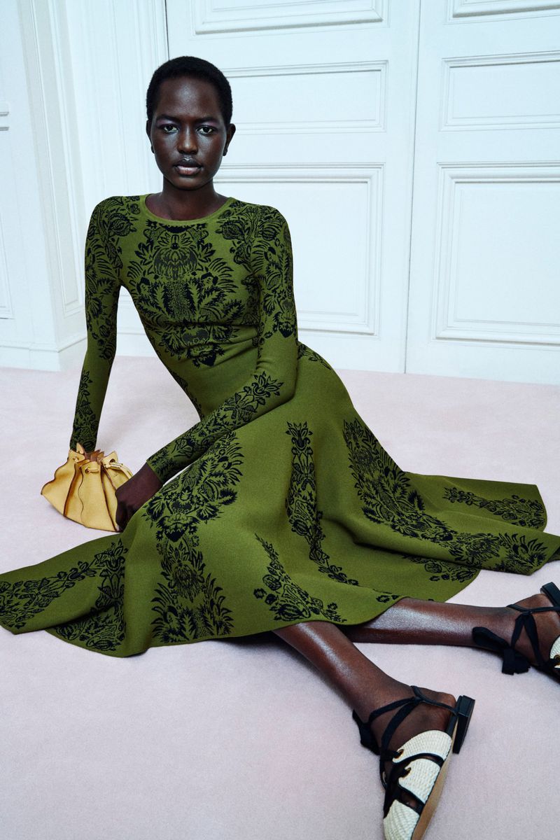 A dark-skinned woman in a long-sleeve olive-green dress with black paisley patterns poses indoors with a small yellow purse.