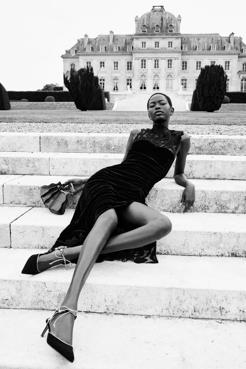 Elegant woman in black velvet dress and heels poses on stone steps in front of a grand European palace on a cloudy day.