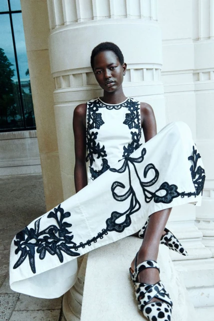 Elegant dark-skinned woman poses in a sleeveless white top and wide-leg pants with black floral embroidery, accented by stylish black and white mules.