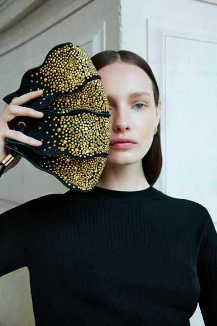 A woman with long brown hair wears a black ribbed sweater and holds a black jeweled clutch over half of her face in a studio setting.