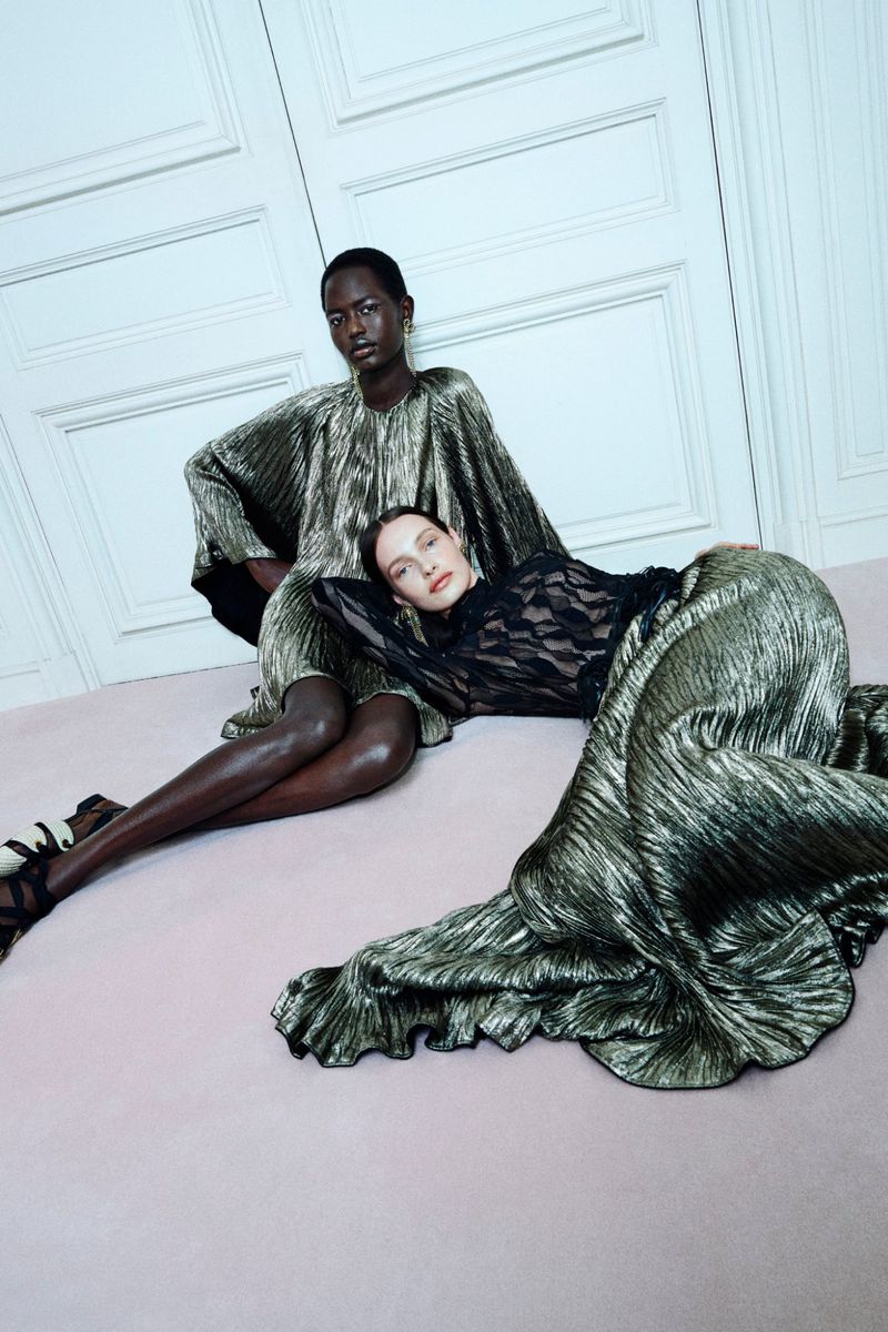 Two women in shimmering metallic dresses pose indoors. One is seated, the other reclining on the floor, creating a fashion-forward composition.