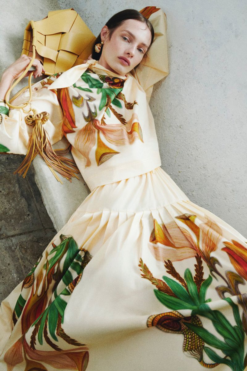 A fair-skinned woman in a floral-print tiered dress reclines against a stone surface, accessorized with a tan woven purse and gold earrings.