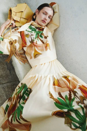 A fair-skinned woman in a floral-print tiered dress reclines against a stone surface, accessorized with a tan woven purse and gold earrings.