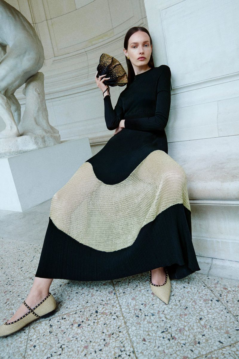 Elegant woman in black top and color-blocked pleated skirt sitting and holding a gold decorative purse in an indoor setting.
