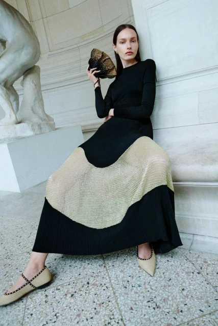 Elegant woman in black top and color-blocked pleated skirt sitting and holding a gold decorative purse in an indoor setting.