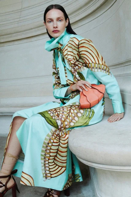 Fashion shot of a woman in an patterned dress sitting in front of a stone wall holding an orange clutch bag looking at the camera.