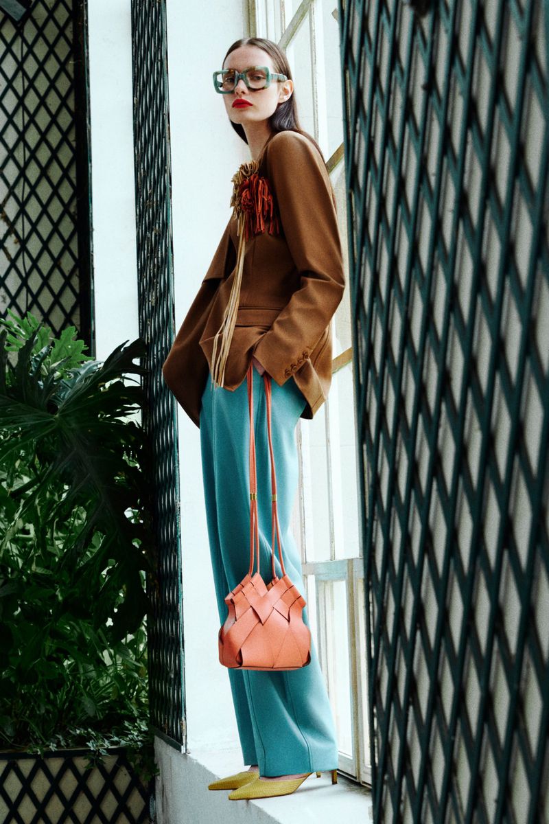A woman poses next to a window, wearing a brown blazer, blue pants, yellow heels, and carrying a peach-colored purse for a fashion editorial.