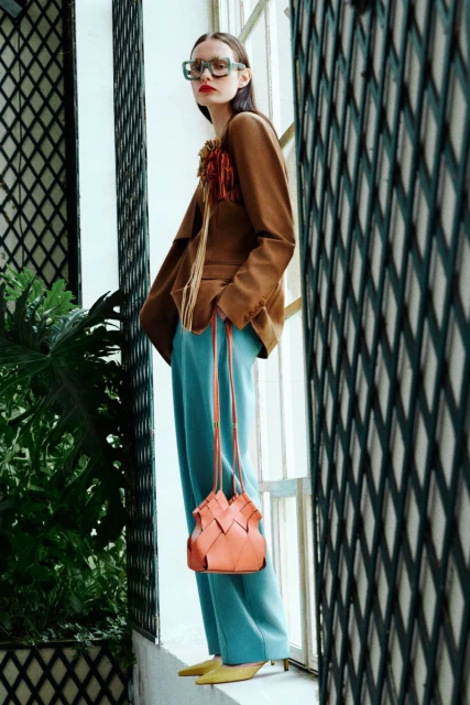 A woman poses next to a window, wearing a brown blazer, blue pants, yellow heels, and carrying a peach-colored purse for a fashion editorial.