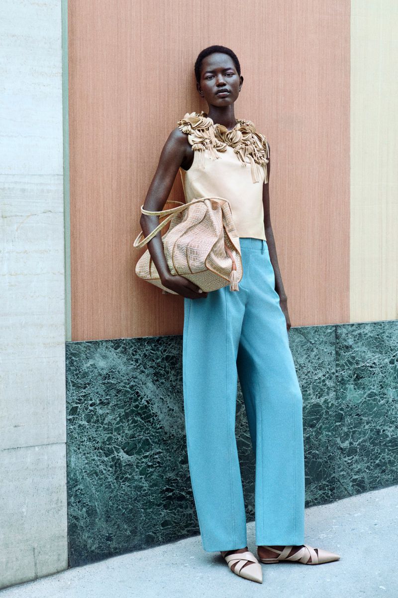 Elegant woman posing outdoors in wide-leg pants and statement top, carrying a handbag; fashion editorial style, full shot.