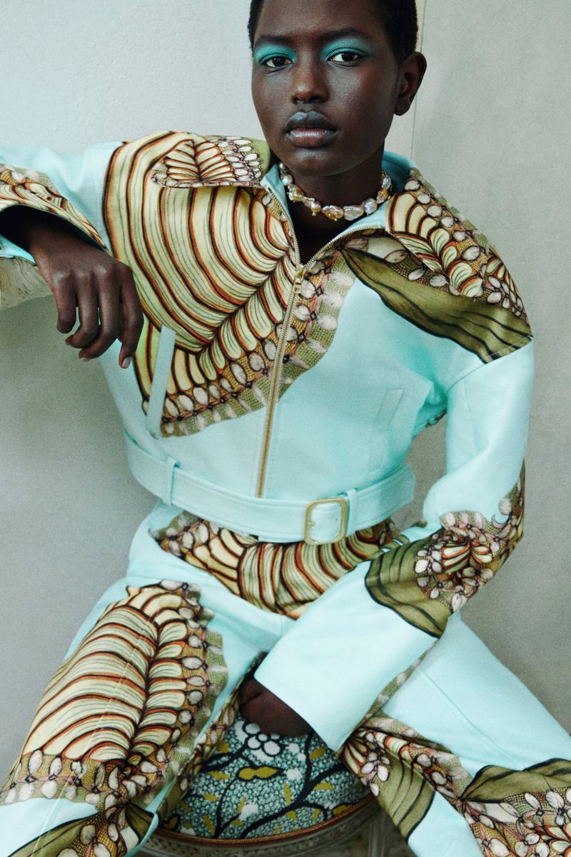 A striking fashion portrait showcasing a Black model in a light blue patterned jacket and pants with matching blue eye makeup against a neutral wall.
