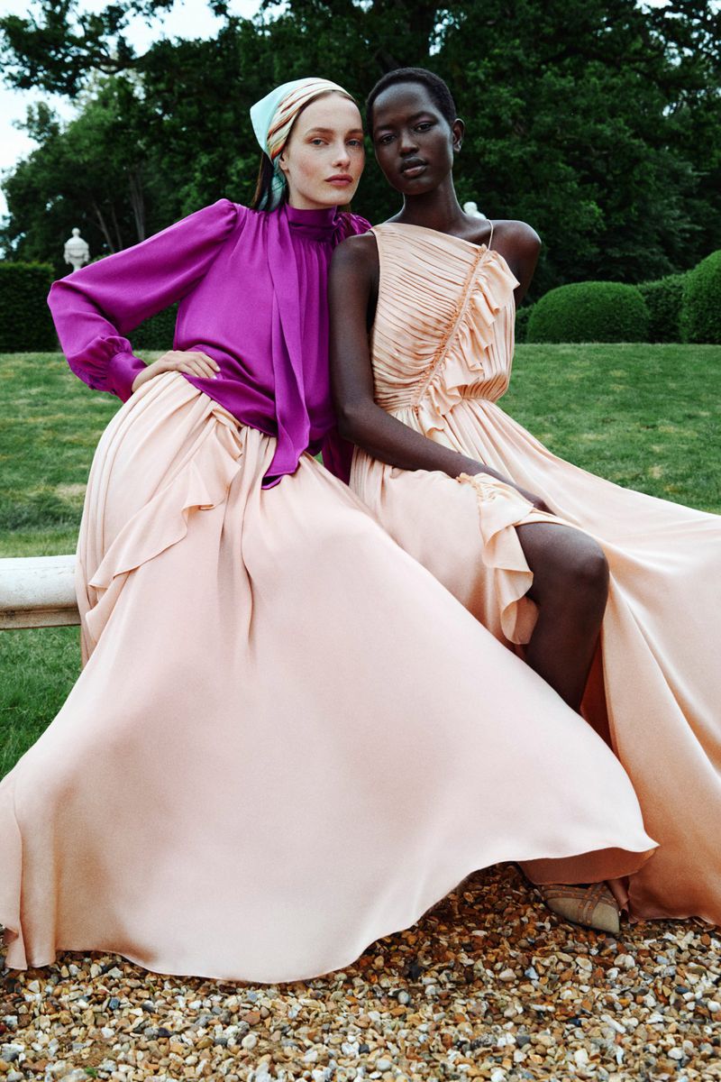 Two fashion models pose in long flowing gowns outdoors. One model wears a purple top with a headscarf and the other a light orange dress.