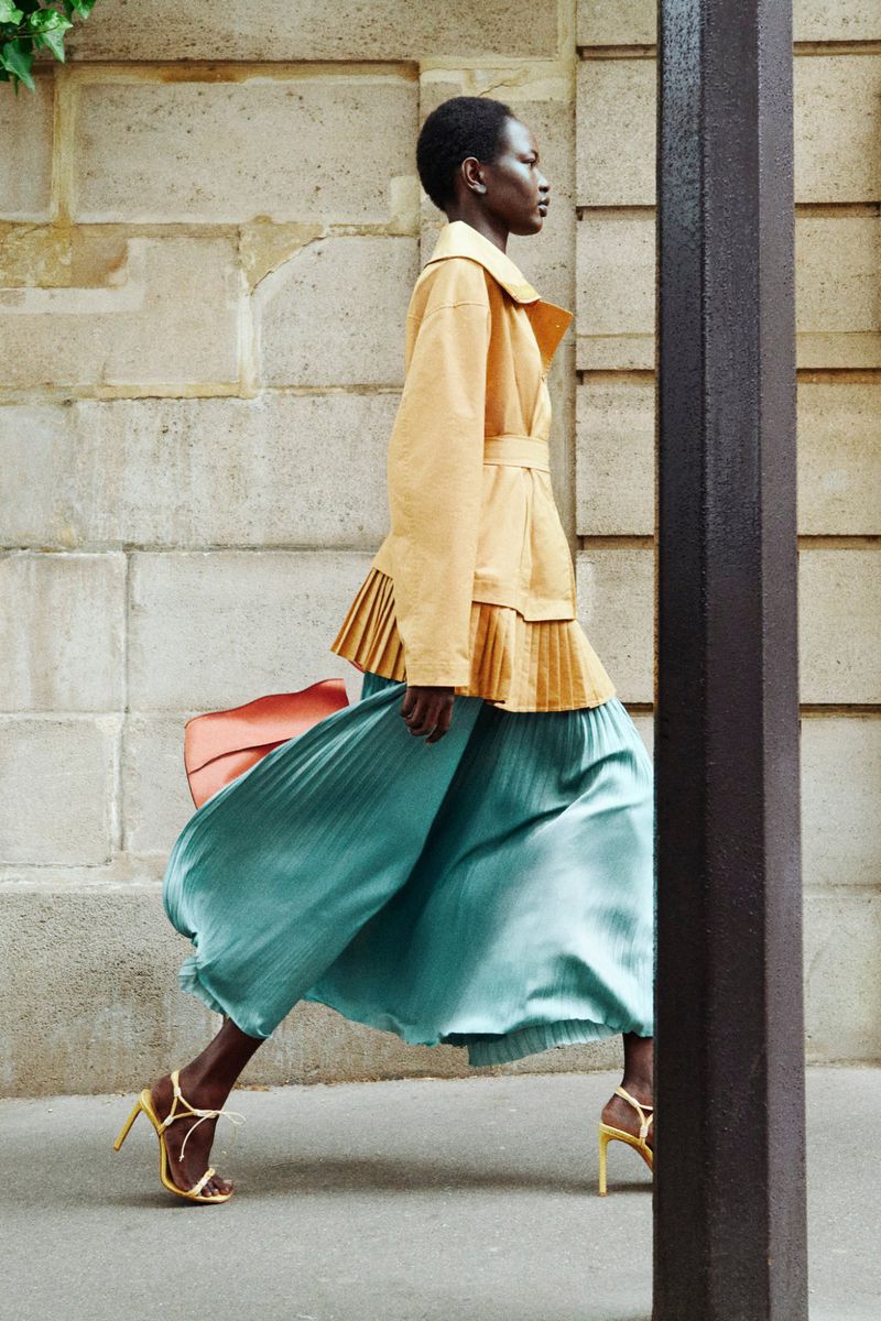An African-American woman walks on a city street wearing a tan jacket, pleated teal skirt, strappy heels and carrying a purse.
