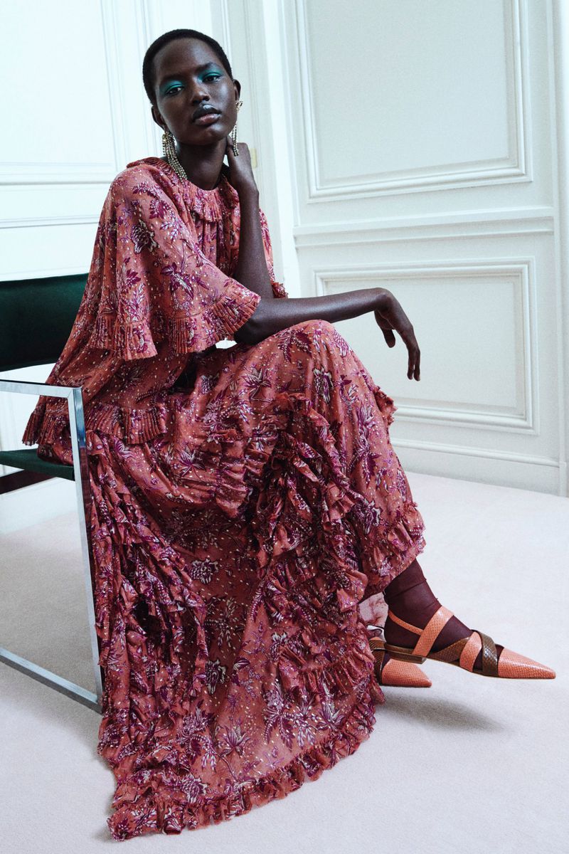 A Black female model poses seated in a flowing pink-patterned maxi dress with ruffles, accessorized with earrings, in a minimalist interior.