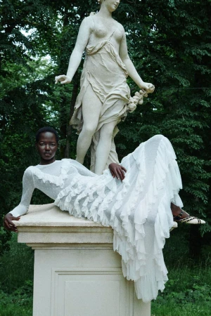 Elegant woman in a white ruffled dress reclines on a pedestal, statuesque, in an outdoor garden setting, trees in the background.