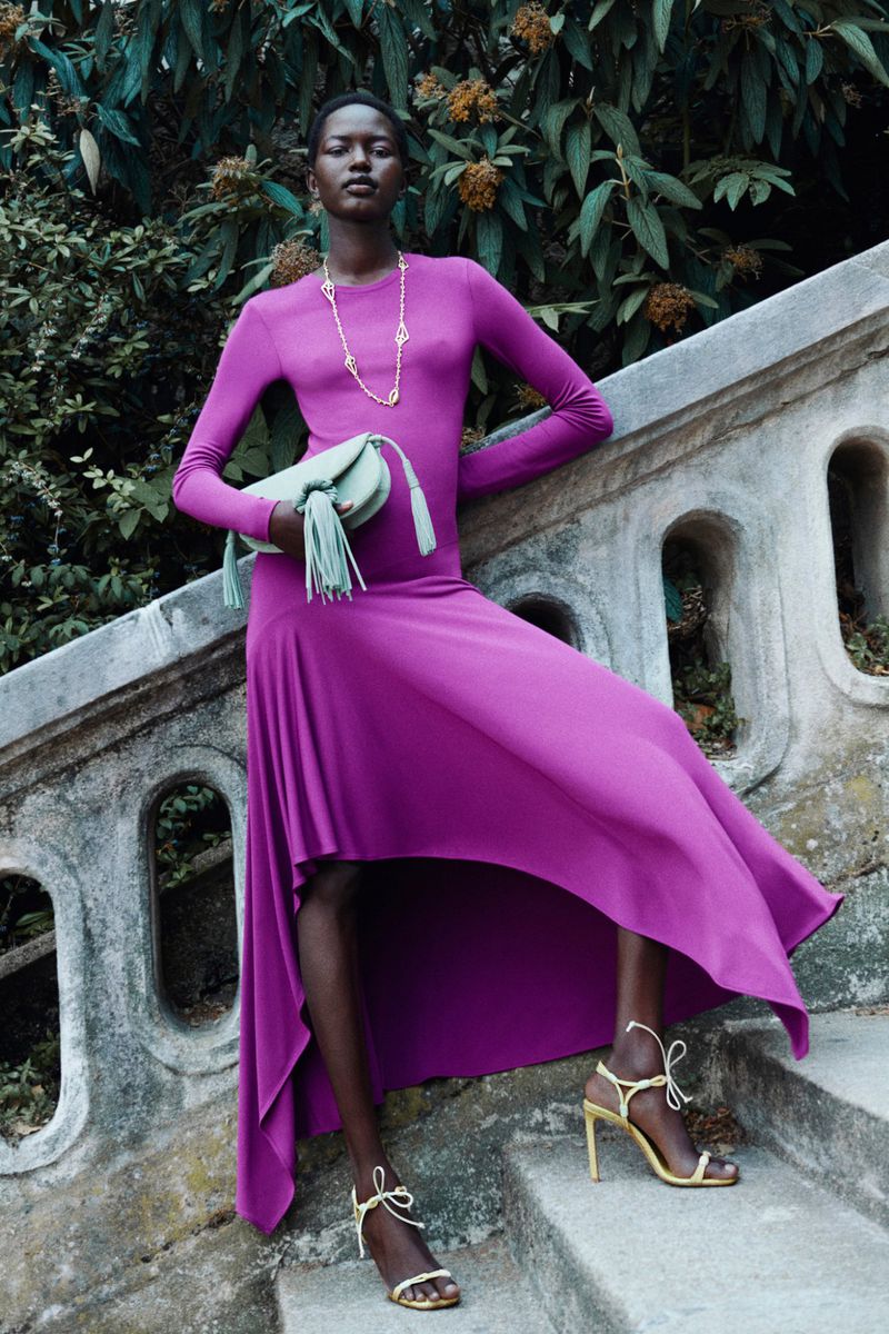 Fashion shot of a slender Black woman in a magenta long-sleeve dress, holding a light-green tassel purse on stairs with green foliage.