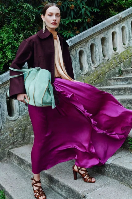 A woman in a maroon jacket and flowing magenta skirt poses on stone steps, carrying a light green fringed purse outdoors.