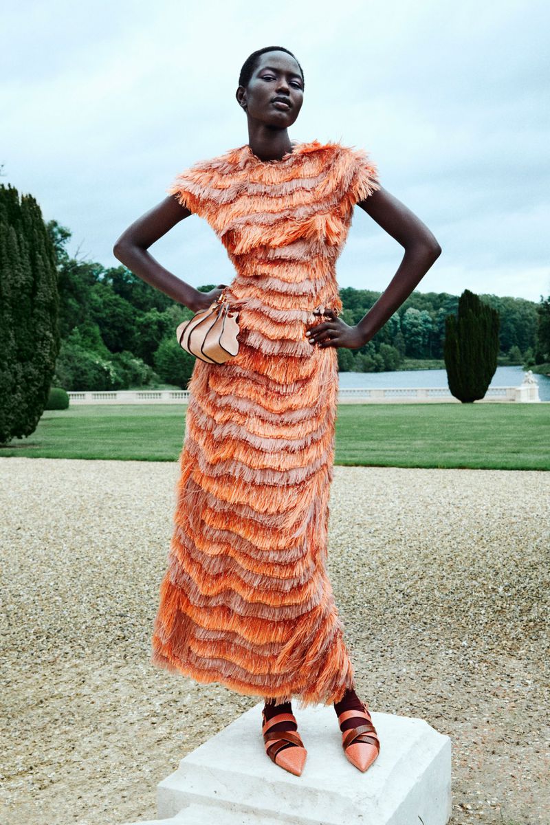A slender woman poses in an orange fringed dress with a purse outdoors on a sunny day against a backdrop of a lawn and trees.