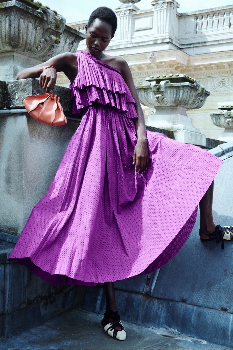 Woman in ruffled one-shoulder dress holding purse. Purple midi dress with polka dots, on rooftop, outdoors with natural light.