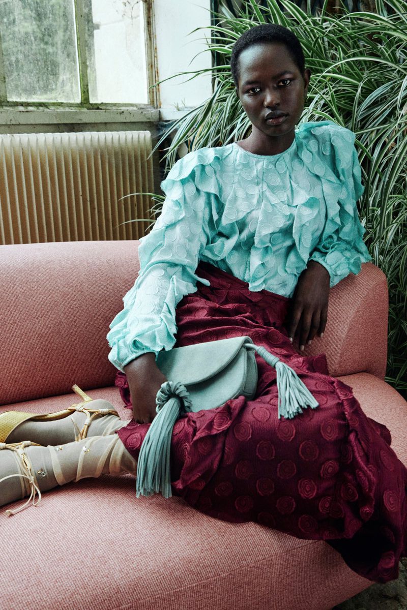 A Black woman sits on a pink sofa in a ruffled turquoise blouse, a burgundy skirt, and tan strappy heels, holding a tasseled purse.