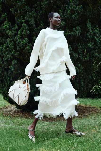 An adult female stands in a grassy outdoor setting, wearing a white fringe sweater and skirt, holding a cream tote bag, and smiling.
