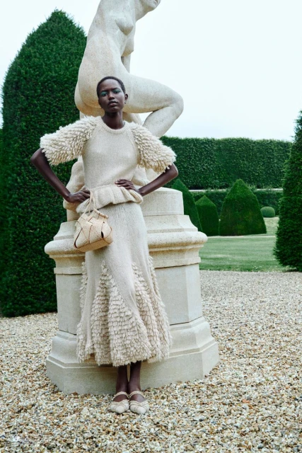 A woman poses in a knit sweater and skirt in a garden setting, beside a stone sculpture and manicured hedges with a gravel walkway.