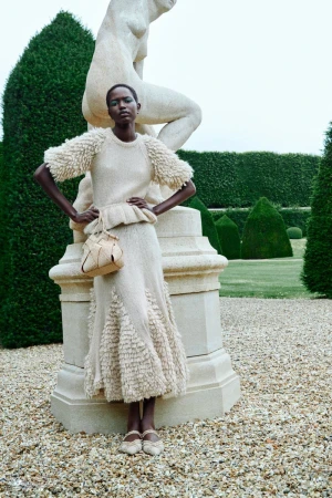 A woman poses in a knit sweater and skirt in a garden setting, beside a stone sculpture and manicured hedges with a gravel walkway.