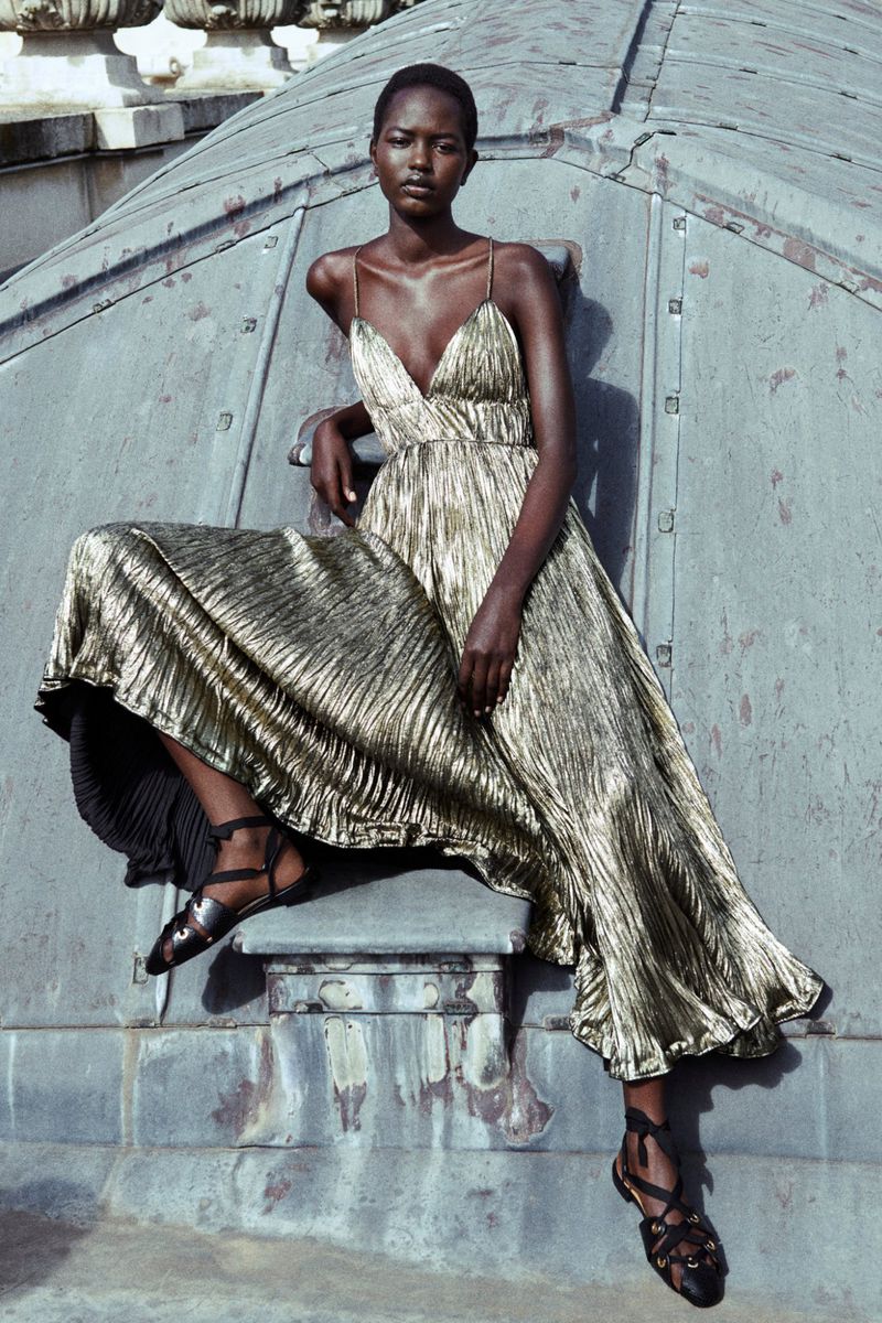 Elegant African-American woman in a shimmering gold dress poses confidently on a rooftop with textured metal elements and simple black sandals.