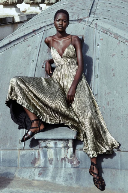 Elegant African-American woman in a shimmering gold dress poses confidently on a rooftop with textured metal elements and simple black sandals.