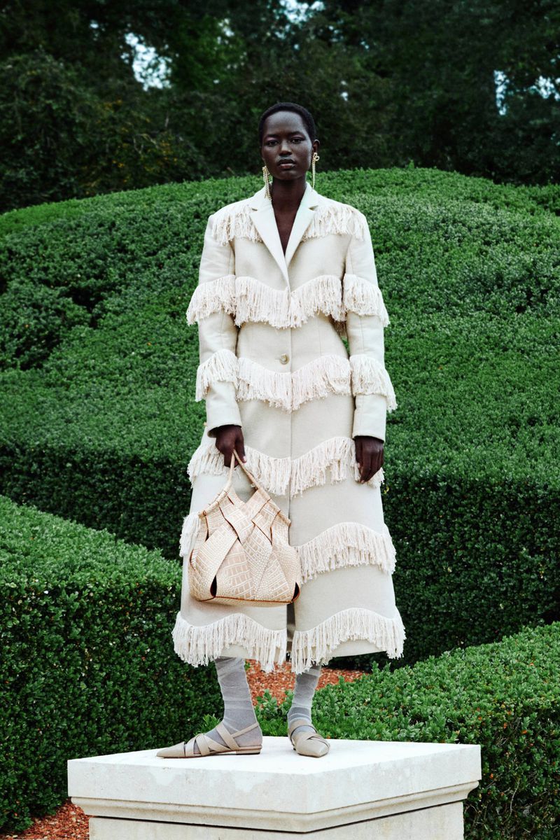 A tall, dark-skinned woman stands on a plinth in an off-white fringe coat, holding a woven bag, in front of sculpted green foliage.