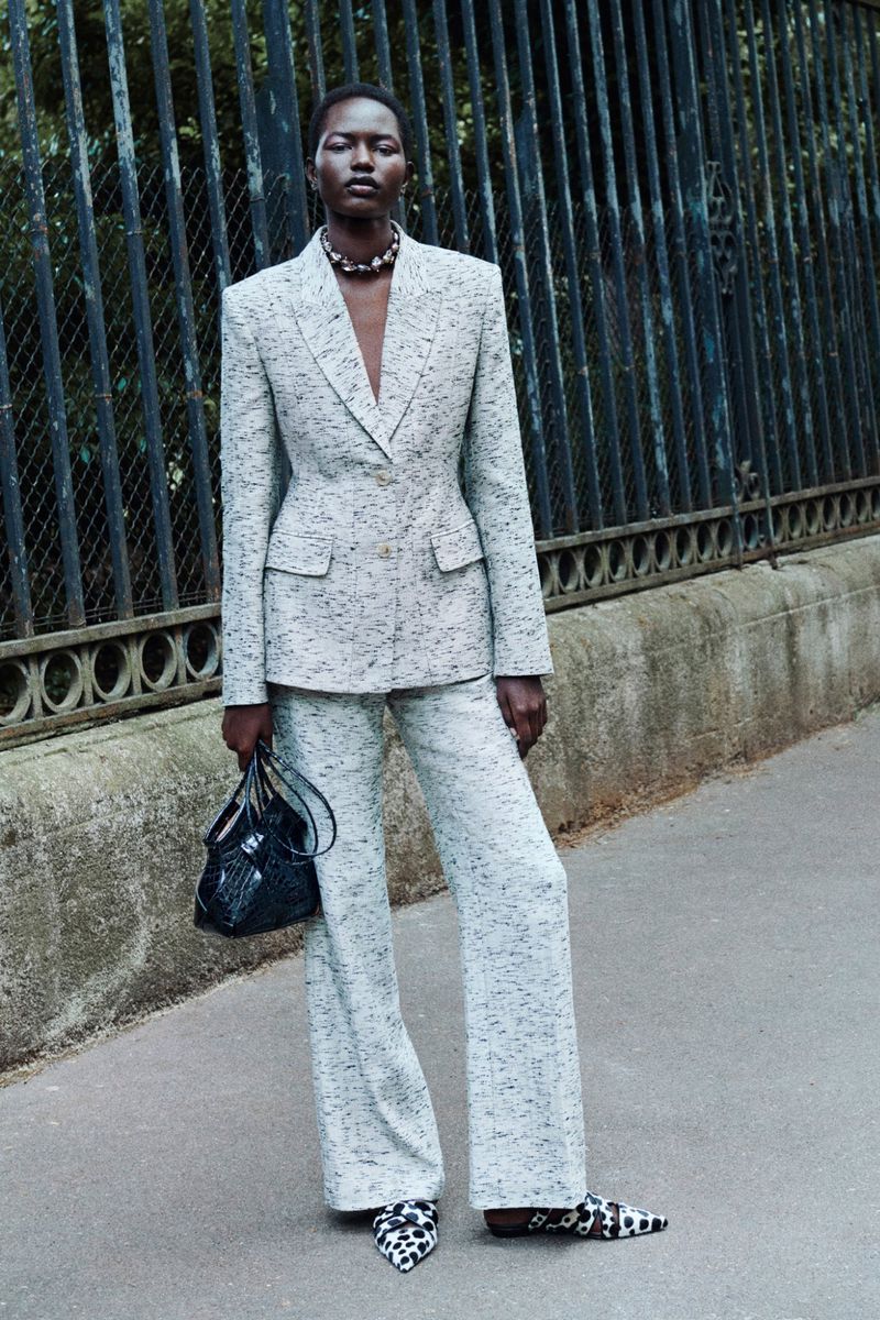 Elegant Black woman in a speckled grey suit and patterned shoes poses outdoors with a black purse, next to a dark metal fence.